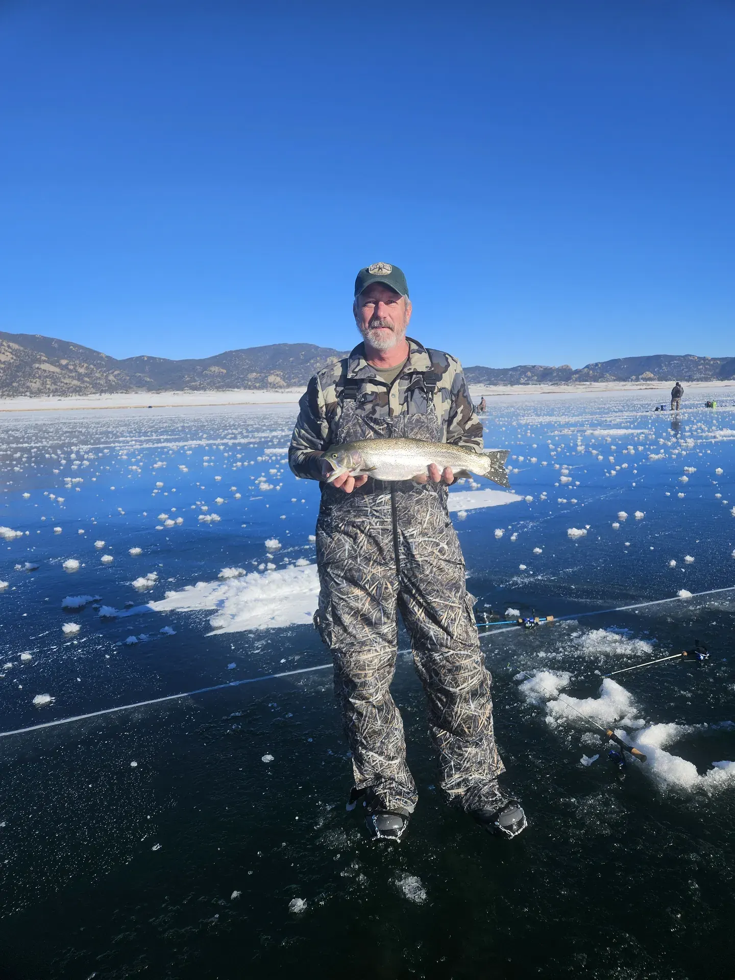 An angler standing on clear blue ice with his catch, mountains along the far shore