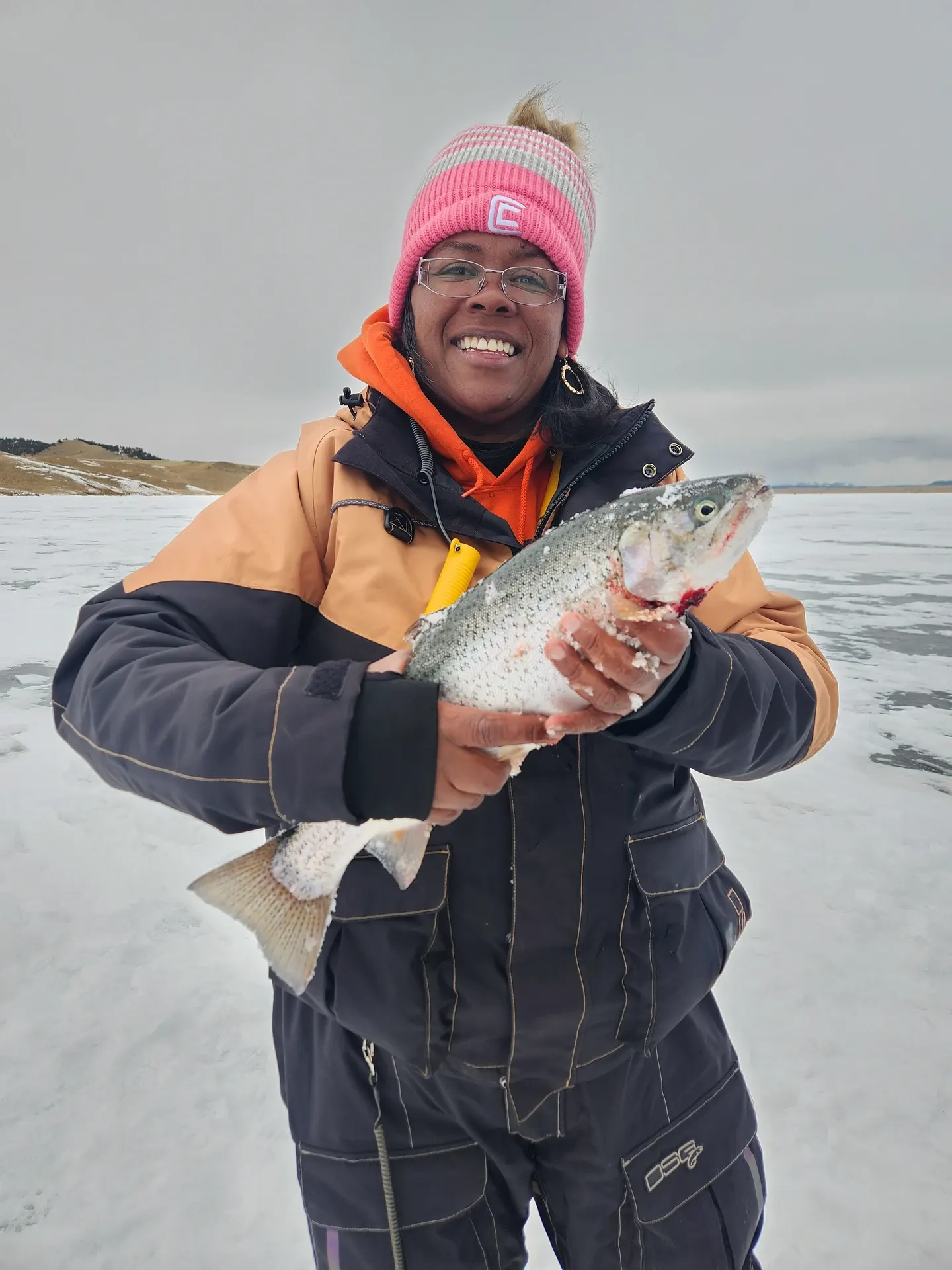An angler in a winter parka and knit hat holding a trout on the ice